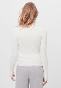 Kobieta z falującymi, jasnobrązowymi włosami, ubrana w długi rękaw, biały sweter prążkowany i jasnoszare spodnie, zwrócona plecami na białym tle.