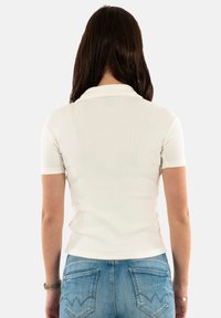 Úzka polo košeľa s krátkym rukávom v off-white farbe, s textúrovanou rebrovanou tkaninou a klasickým golierom. Spárovaná s modrými džínsami.