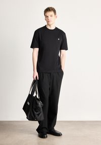 Μαύρο κοντομάνικο μπλουζάκι με στρογγυλή λαιμόκοψη και μικρό λογότυπο. Συνδυασμένο με φαρδιά μαύρη παντελόνια και κρατώντας μαύρη τσάντα με φερμουάρ.