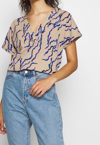 Béžová blůza s krátkým rukávem a hlubokým výstřihem do V, s vlnitými modrými a růžovými vzory, spárovaná s světle modrými džíny s vysokým pasem.