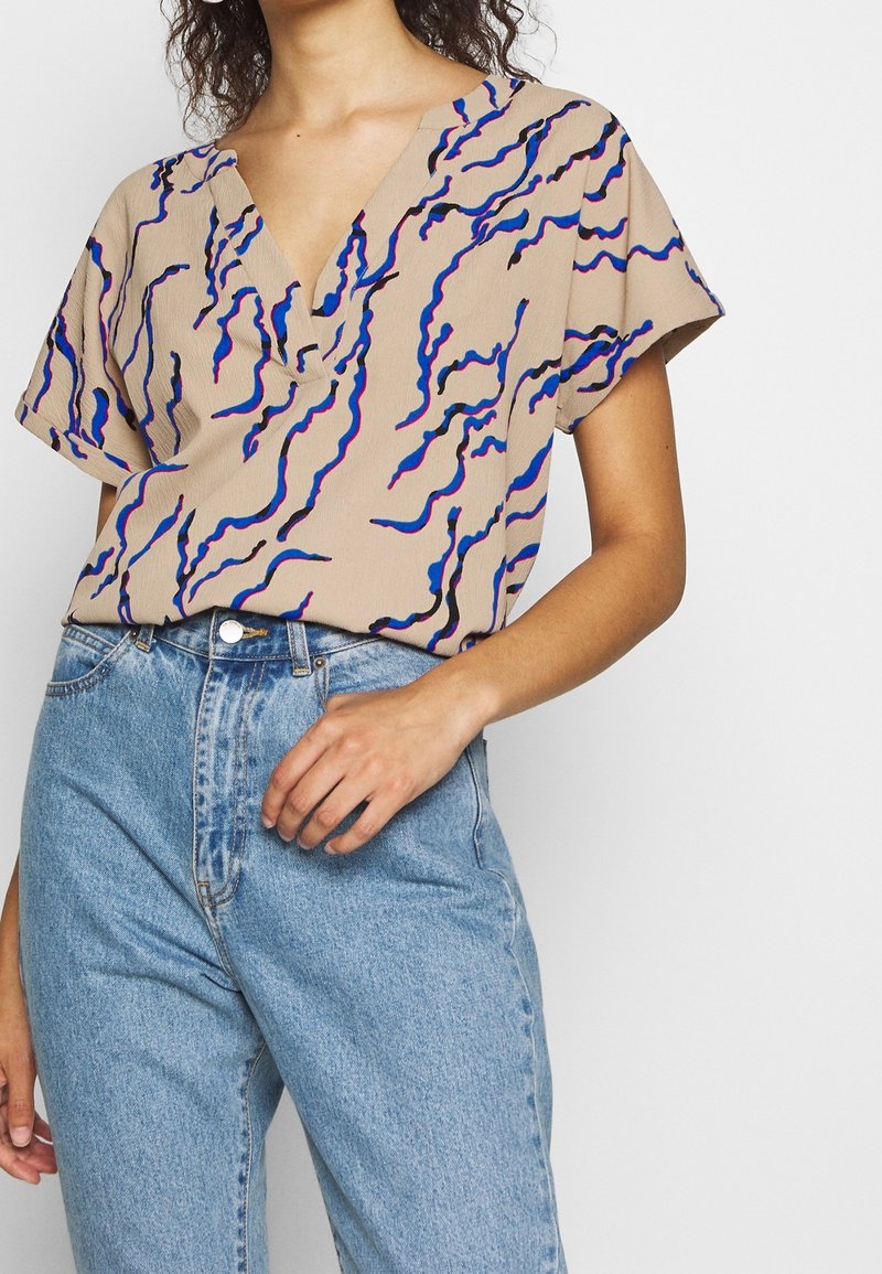 Béžová blůza s krátkým rukávem a hlubokým výstřihem do V, s vlnitými modrými a růžovými vzory, spárovaná s světle modrými džíny s vysokým pasem.