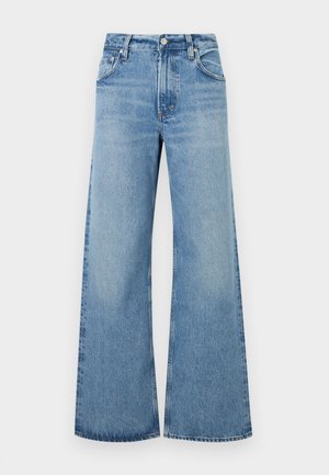 Žena oblečená v čierno-bielom pruhovanom tričku s krátkym rukávom, svetlomodré džínsové nohavice so širokými nohavicami, čierne sandále na podpätku, drží pod pazuchou čiernu koženú tašku.