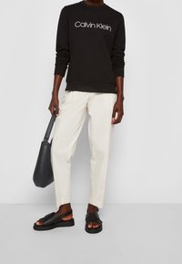 Zwarte sweatshirt met witte "Calvin Klein" logo, crèmekleurige broek, zwarte sandalen met kruisbandontwerp en een zwarte leren schoudertas.