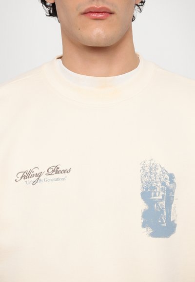 Beiger Sweatshirt mit Rundhalsausschnitt und gerippten Bündchen. Mit einem Frontdruck in braunem und blauen Text sowie einer Illustration einer Person und Blumen.