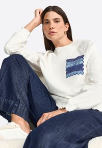 Felpa bianca con patch ricamato blu caratterizzato da motivi ondulati; indossata con jeans a gamba larga blu scuro; vestibilità rilassata e polsini a costine.