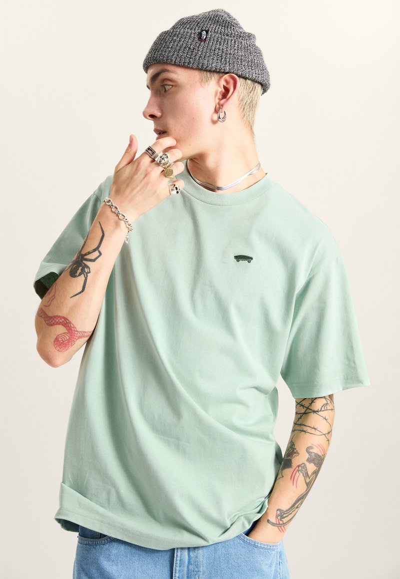 Mintgrøn kortærmet T-shirt med et skateboardlogo, sammen med en grå strikket hue. Har detaljerede tatoveringer på underarmen.
