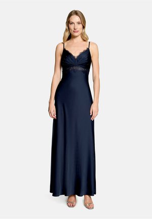 Femme portant une longue robe nuisette bleu marine avec une bordure en dentelle, des bretelles spaghetti et des talons ouverts argentés, debout sur un fond clair uni.