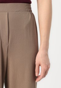 Lichtbruine broek van een gemêleerde stof. Heeft een ingezette elastische tailleband, voorplooien en een soepele silhouet.