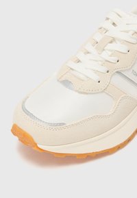 Beige and white sneaker made of suede and mesh, featuring a textured toe cap, rubber outsole, and a lace-up design with silver accents.