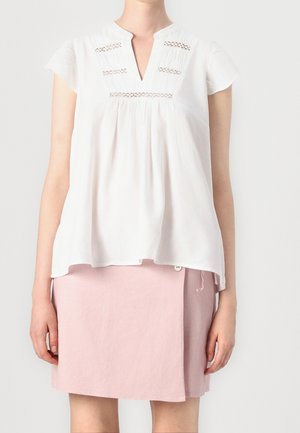 Femme portant une blouse blanche à manches courtes avec des détails en dentelle et une jupe asymétrique rose boutonnée, debout devant un fond uni.