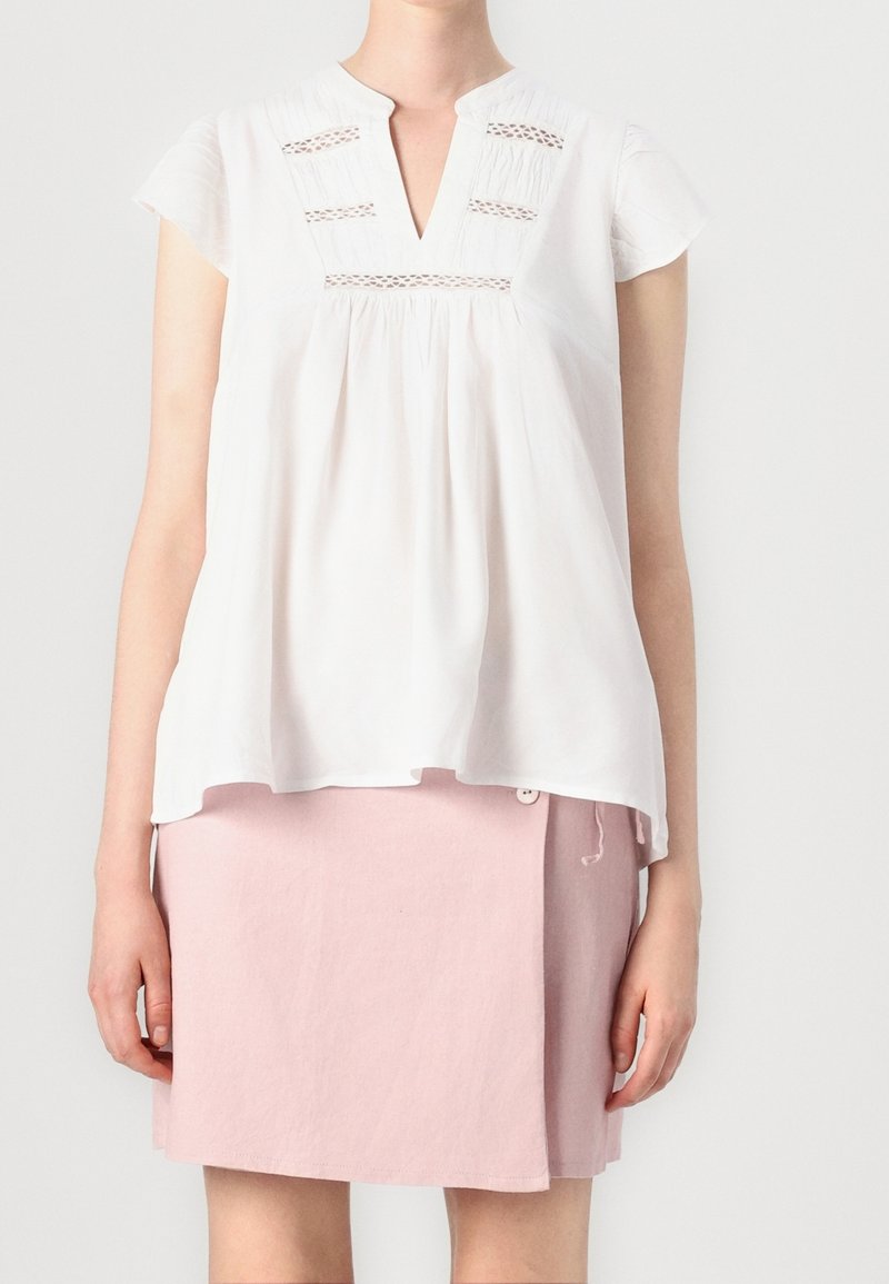 Femme portant une blouse blanche à manches courtes avec des détails en dentelle et une jupe asymétrique rose boutonnée, debout devant un fond uni.