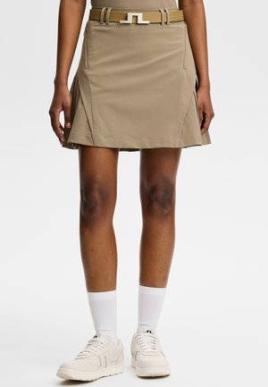 Persoon die een beige rok draagt met bijpassende riem, witte sokken tot halverwege de kuit en witte vetersneakers tegen een effen achtergrond.