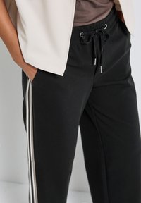 Personne portant un pantalon noir à cordon avec des rayures blanches sur les côtés, une veste beige et un haut marron, la main dans la poche sur fond uni.