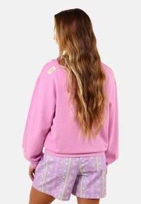 Sweatshirt rose à manches longues, en tissu texturé, avec une petite étiquette logo. Associé à un short violet clair affichant des motifs géométriques.