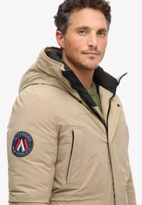 Chaqueta aislante beige con capucha desmontable, acentos en negro, bolsillos con cierre y un parche circular con logo en la manga izquierda.