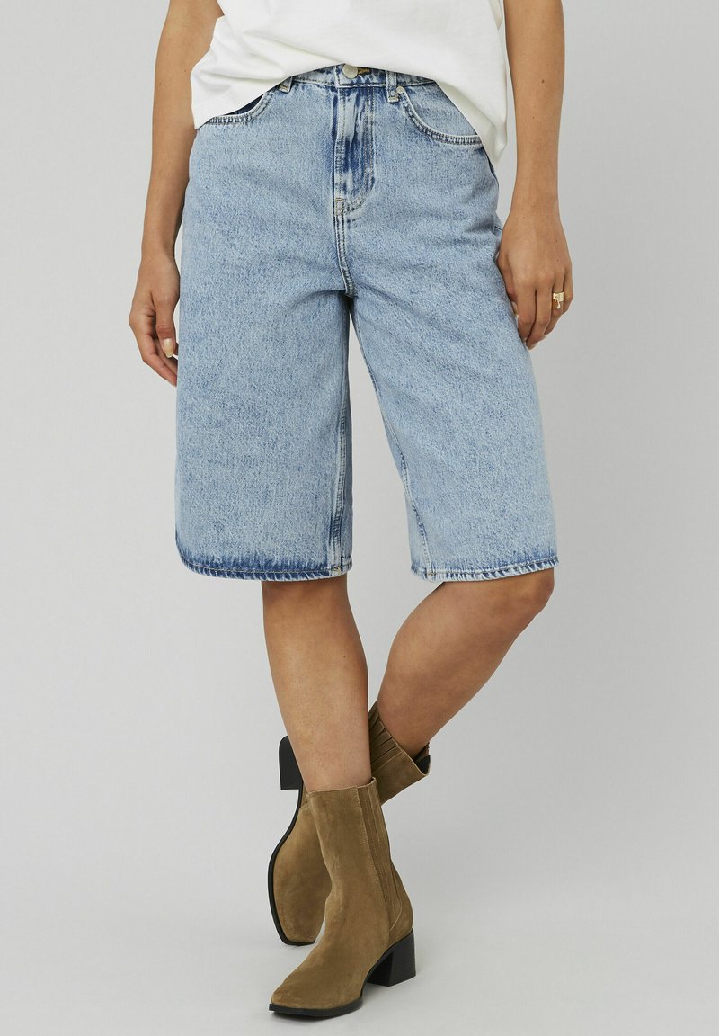 Un short en denim bleu clair, longueur Bermuda, porté avec des bottines en daim beige et un haut blanc rentré, visible de mi-thigh à mi-mollet.