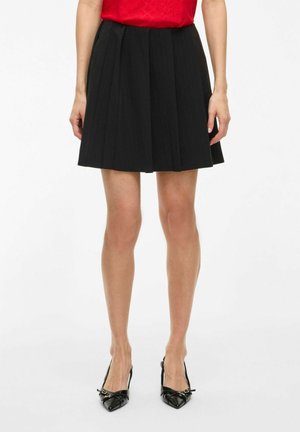 Jupe plissée noire avec une taille ajustée, fabriquée à partir d'un tissu lisse. Associée à des escarpins noirs à bout pointu ornés d'accents décoratifs.