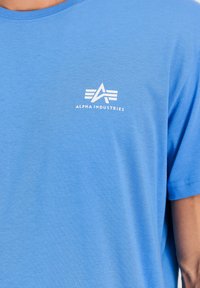 Zila īsām piedurknēm T-krekls, kas izgatavots no mīksta auduma, ar baltu logotipu un grafisko dizainu augšējā kreisajā krūšu zonā.