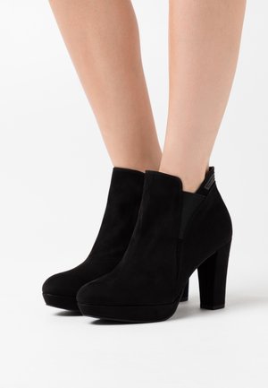 Botines de ante negro con tacones cuadrados, llevados sobre piernas al desnudo contra un fondo blanco.