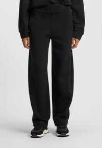 Svarta sweatpants i mjukt material, med avslappnad passform, elastisk midja och smalnande ben, tillsammans med svarta sneakers.