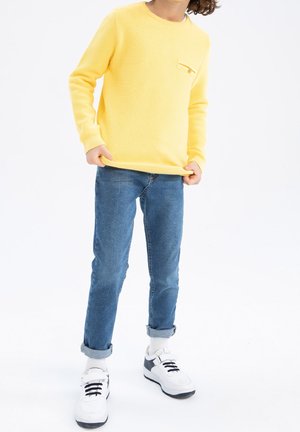 Niño con suéter amarillo de manga larga, vaqueros azules remangados, calcetines blancos y zapatillas blancas, de pie con las manos sujetando el borde del suéter.