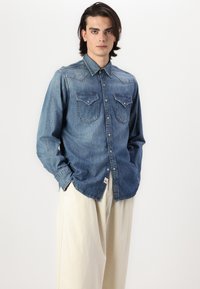 Camicia di jeans azzurro chiaro con taglio classico, caratterizzata da due tasche sul petto con pattine a bottone, colletto a punta e sottili dettagli di cucitura.
