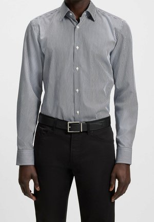 Hombre que lleva una camisa abotonada con rayas verticales en blanco y negro metida en pantalones negros con un cinturón negro, sobre un fondo liso.