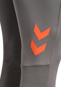 Close-up van grijze sportlegging met twee feloranje chevronpijlen op de dij en getextureerde kniekussens.