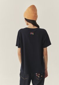 Tumesinine puuvillane t-särk lõdva lõikega, mille tagaküljel on värvikas tikkimistekst. Sobitatud soonikkoega oranži kootud mütsiga.