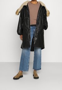 Cappotto di pelle nera con cappuccio foderato in pelliccia, due tasche frontali e chiusura con zip. Indossato sopra un top marrone, abbinato a jeans a gamba larga e scarpe color cammello.