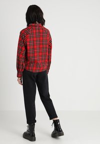 Chemise en flanelle à carreaux rouge avec manches longues et col, portée avec un pantalon noir fuselé et des bottes noires à lacets. Vue de dos visible.