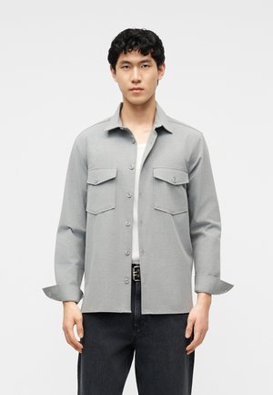 Mężczyzna ubrany w jasnoszarą kurtkę zapinaną na guziki z dwoma kieszeniami na piersiach, białą podkoszulkę, czarny pasek i ciemne spodnie, stojący na jednolitym tle.