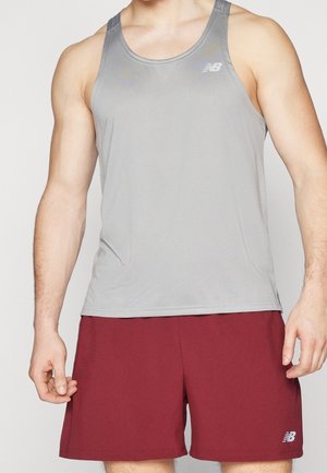 Man draagt een lichtgrijs mouwloos sportshirt en donkerrode korte broek, staat met ontspannen armen langs het lichaam tegen een effen achtergrond.