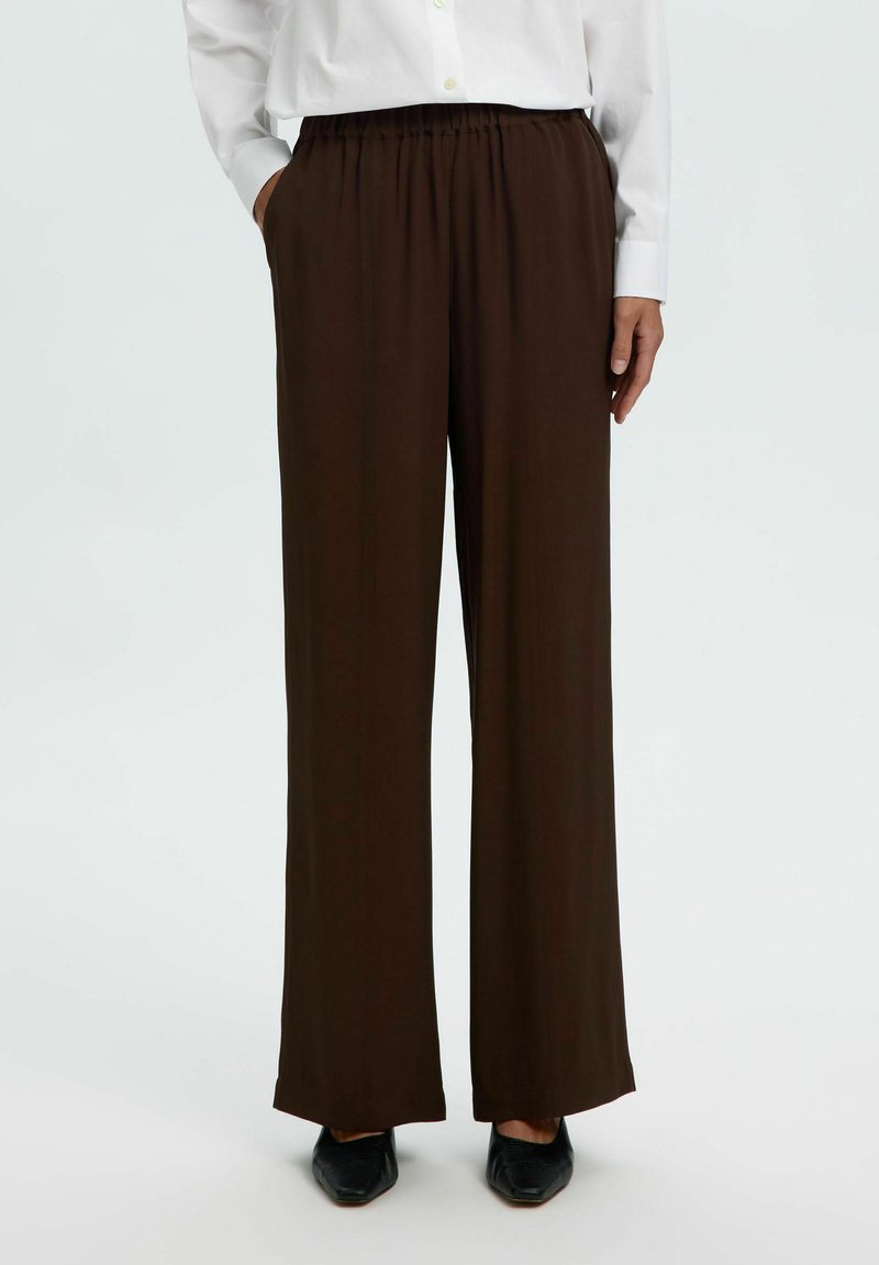 Personne portant un pantalon marron foncé taille haute à jambes larges avec poches latérales, une chemise blanche boutonnée et des chaussures noires pointues.