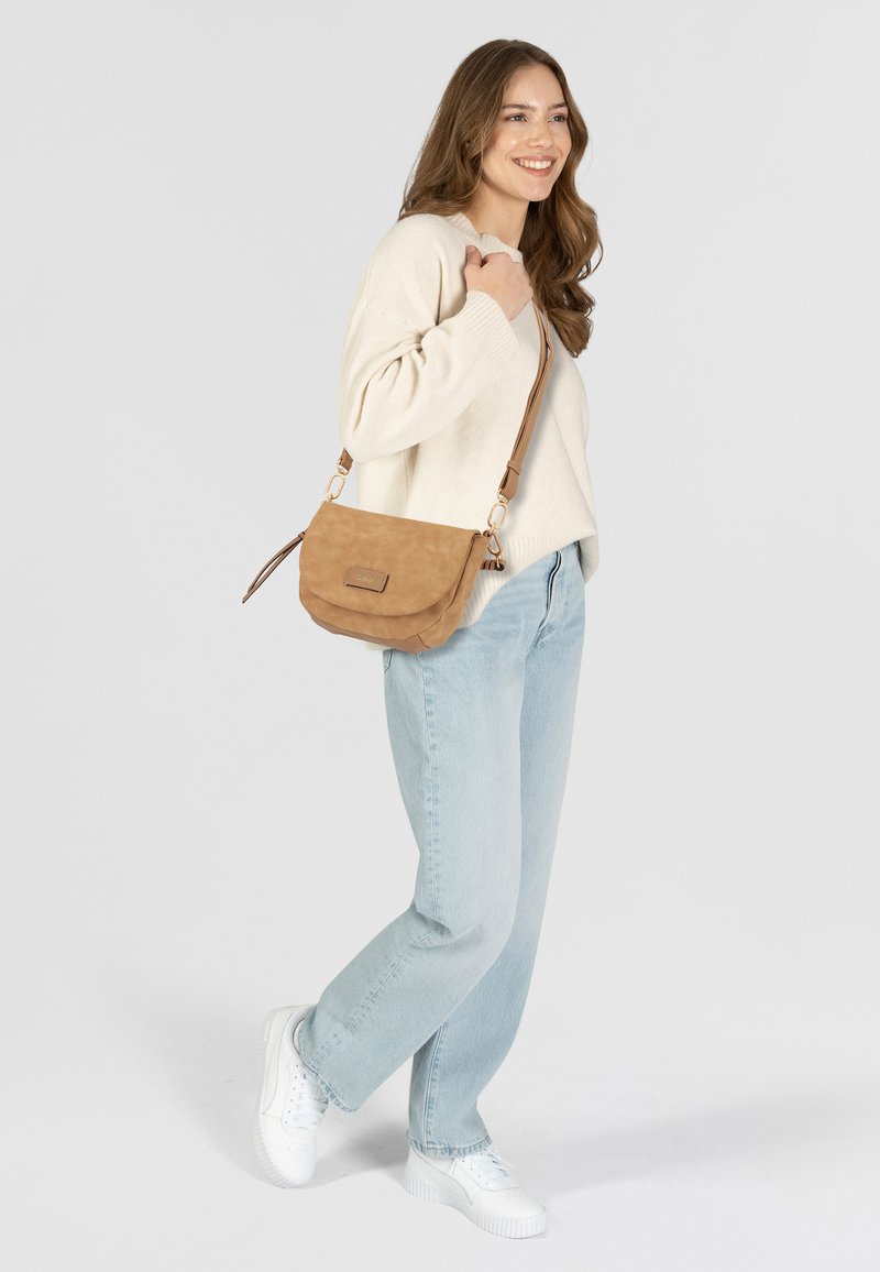 Tasche mit Cross-Body-Trageweise in Tan mit weicher Textur, Klappenverschluss und verstellbarem Träger, kombiniert mit hellblauen Jeans und einem cremefarbenen Pullover.