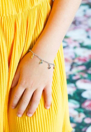 Silbernes Armband mit Charms in Form von Sternen und Herzen, getragen an einer Hand neben einem gelben, plissierten Kleid. Glatte Metallringe und farbenfrohe Charms.