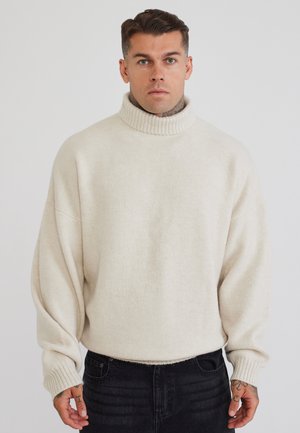 Mand med kort brunt hår og tatoveringer iført en cremefarvet rullekravesweater og sorte jeans, stående mod en ensfarvet baggrund.
