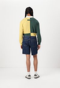 Color-blocked long-sleeve shirt in yellow and green, paired with dark denim shorts. Casual footwear completes the outfit. Minimalist design.m