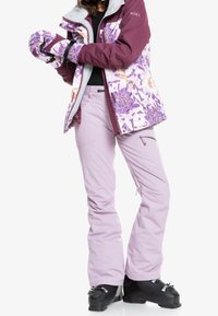 Kvinna i lila blommig skidjacka, lavendelfärgade snöbyxor, vantar och svarta skidstövlar, som justerar sin handske.