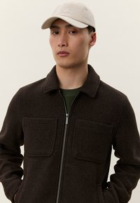 Ruda vilnos mišinio striukė su apykakle, priekiniais užtrauktukais ir dviem krūtinės kišenėmis. Dėvima ant žalios aprangos, derinama su šviesios spalvos kepure.