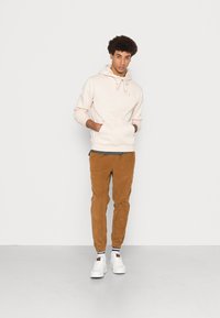 Homem com cabelo encaracolado, vestindo um hoodie creme, calças de veludo cotelê castanhas e tênis brancos, de pé contra um fundo claro liso com as mãos no bolso do hoodie.