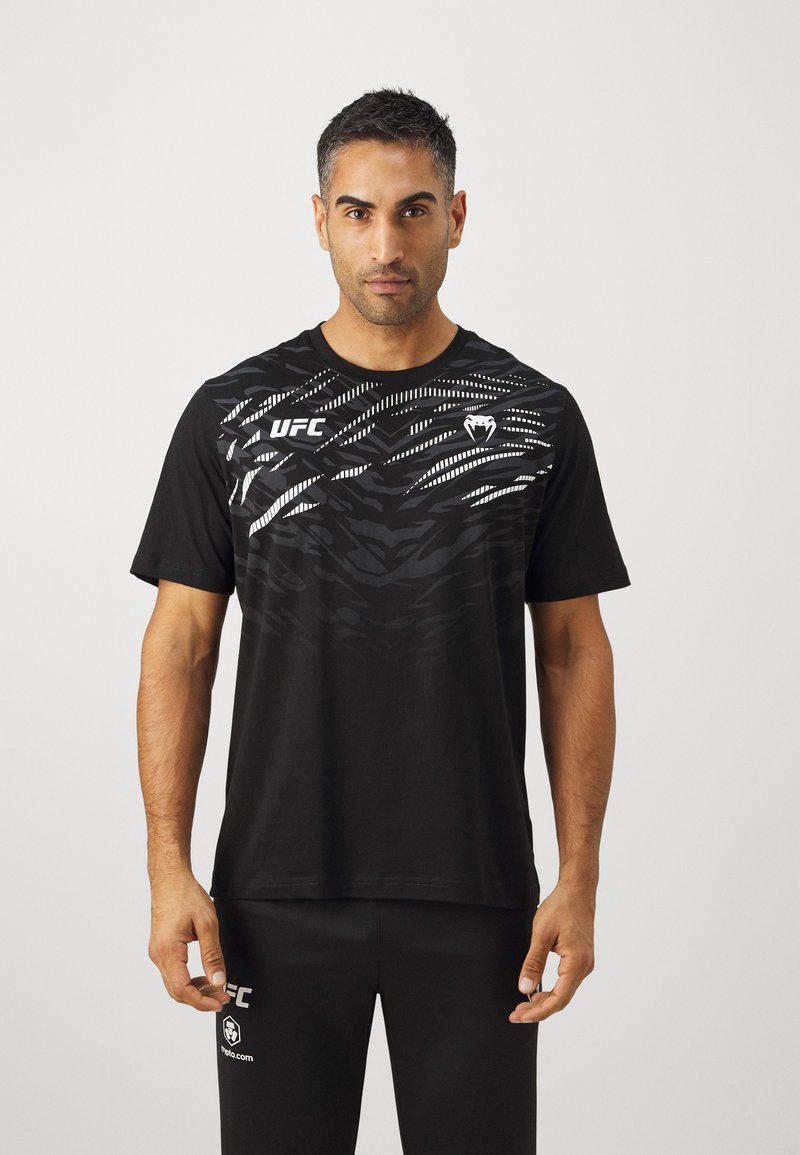 Camiseta negra de manga corta con un atrevido patrón geométrico blanco, y un logo de UFC en el lado izquierdo del pecho. Tela suave con un ajuste relajado.