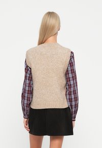 Beige pletená vesta s žebrovaným výstřihem, nošená přes kostkovanou košili s dlouhým rukávem, kombinováno s černou corduroy sukní. Měkká textura, ležérní styl.