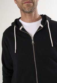 Schwarze Zip-Hoodie mit weißem Kordelzug, ausgestattet mit einem silbernen Reißverschluss und einem darunterliegenden, geschichteten weißen T-Shirt. Aus weichem Stoff gefertigt.