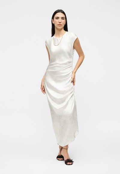 Γυναίκα με μακριά σκούρα μαλλιά, που φορά ένα λευκό αμάνικο σατέν μίντι φόρεμα και μαύρα πέδιλα με δέσιμο στον αστράγαλο, στέκεται με το ένα χέρι στη μέση.