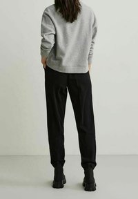 Sudadera gris con cuello redondo, dobladillo acanalado y mangas largas, combinada con pantalones negros y botas de tobillo, de pie sobre un suelo claro.