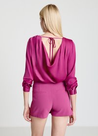 Blouse en satin violet avec un profond décolleté en V dans le dos et un détail de lien, associée à un short assorti sur mesure avec poches latérales. Texture lisse partout.