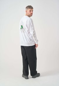 Langärmliges weißes T-Shirt mit einem grünen Fisch-Graphic und Spinnennetzdesign auf dem Rücken, mit Text entlang der Ärmel. Schwarze Hosen und Schuhe.