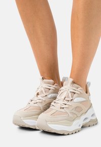 Chaussures de sport beige en mesh et daim, avec semelles blanches épaisses et design à lacets. Accents de logo remarquables sur les côtés et la languette.