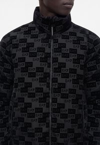 Homme portant une veste noire avec des logos EA7 répétés, une fermeture éclair frontale et un col montant, visible des épaules jusqu’aux lèvres sur un fond uni.